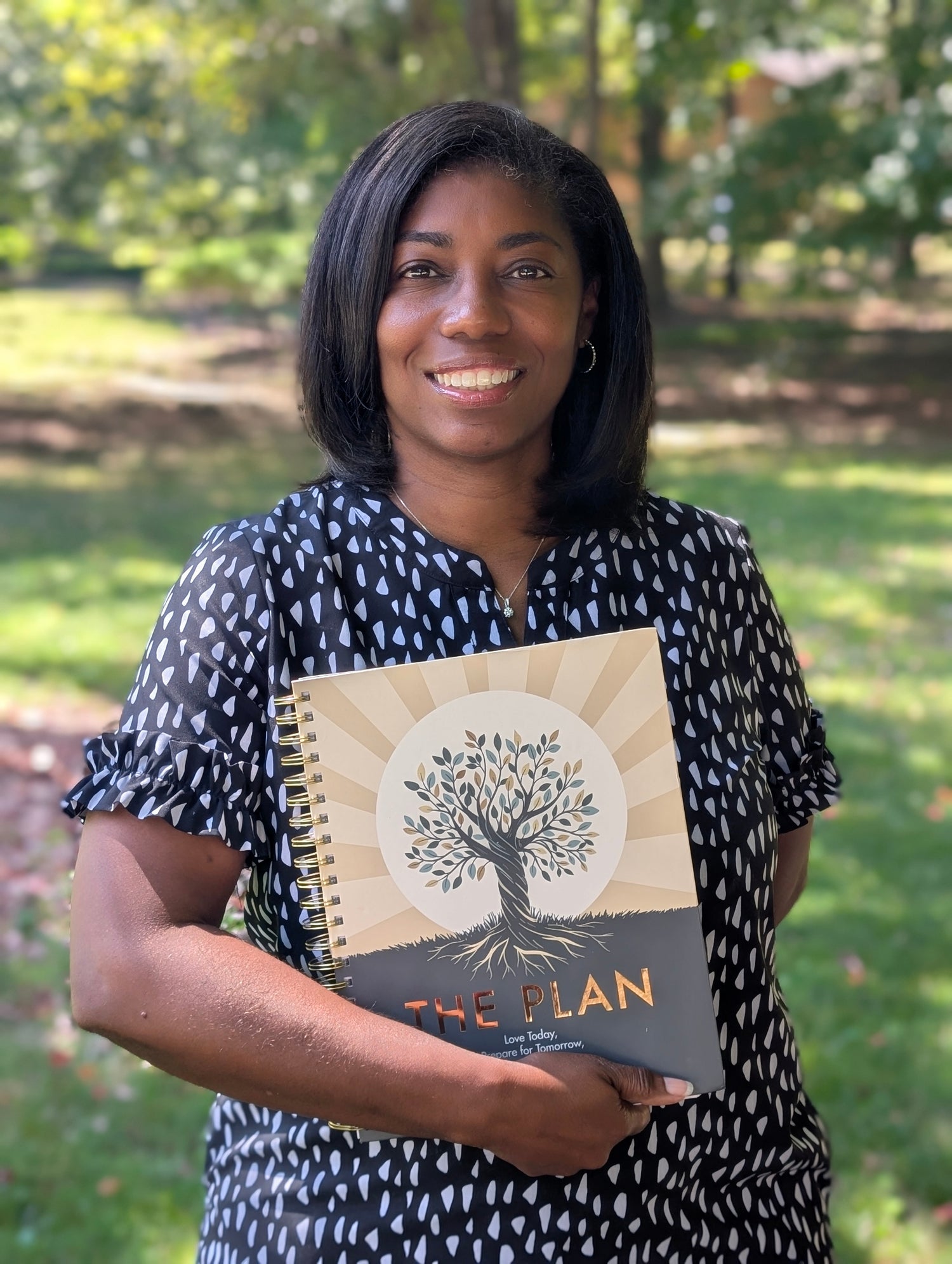 Woman holding a planner titled 'The Plan' outdoors with greenery in the background